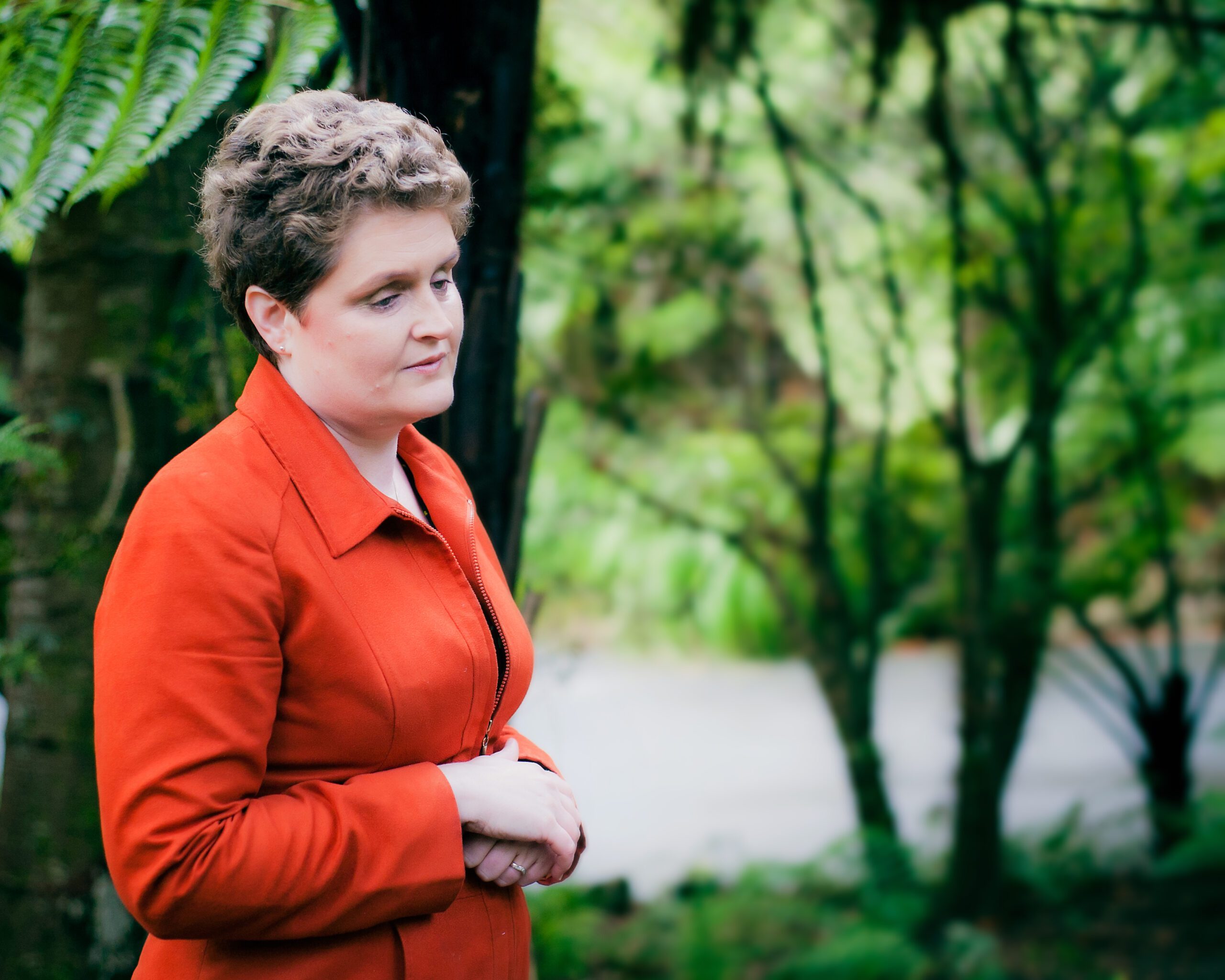Lisette in an orange shirt, holding her hands. There are trees and foliage in the background.